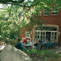 Menschen sitzen im Außenbereich eines Landcafés unter Bäumen und genießen Getränke