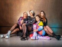 Medlz Vier junge Frauen in 90er Jahre Look sitzen lässig vor einer Wand.